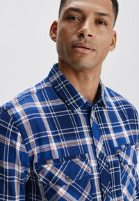 Niebieska koszula w kratę zapinana na guziki, uszyta z tkanej tkaniny; posiada kołnierz, dwie kieszenie na piersiach oraz mieszankę białych i burgundowych pasków.