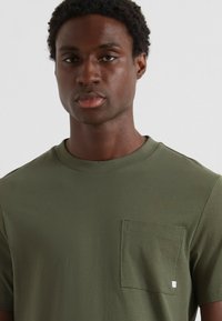 T-shirt vert olive en coton doux, avec un col rond et une poche plaquée sur le côté gauche de la poitrine. Design simple, manches courtes.