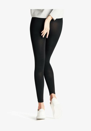 Leggings noirs en tissu extensible, avec une texture lisse et un design ajusté. Associés à des baskets blanches.