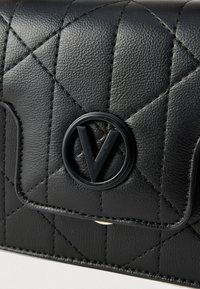 Musta nahkainen lompakko, jossa on tikattu kuvio ja pyöreä musta embleemi, jossa on tyylitelty "V"-logo ja kontrastitikkaukset.