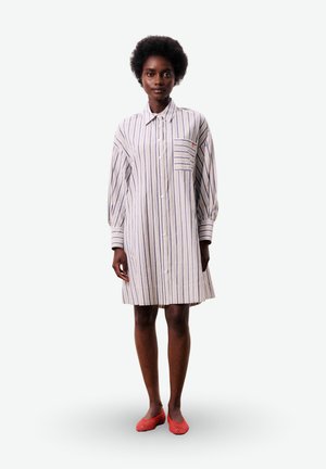 Vrouw staat en draagt een loszittende, knielange gestreepte overhemdjurk met lange mouwen en rode platte schoenen tegen een effen witte achtergrond.