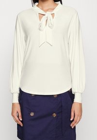 Krémová blůza s mašlí na krku, dlouhými nafouknutými rukávy a otvorovým výstřihem. Hladká tkanina s jemným vzhledem.