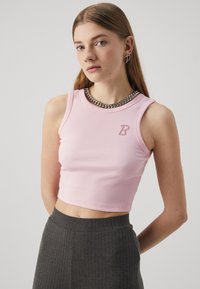 Ružový pletený crop top s okrúhlym výstrihom a malým logom "B" vykladaným krištáľmi. V páre s tmavošedými plisovanými nohavicami s vysokým pásom.