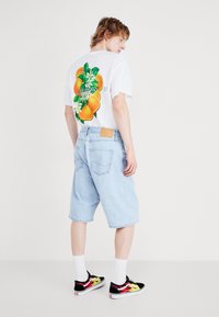 Hvit T-skjorte med oransje og blomstergrafikk på ryggen, sammen med lyseblå denimshorts og svarte joggesko med flammeaksenter.