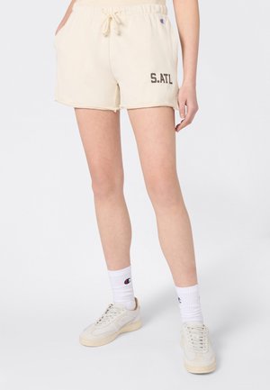 Kobieta w beżowych szortach na sznurek z napisem „S. ATL”, białych skarpetkach Champion i beżowych sneakersach, stojąca na tle jednolitego tła.