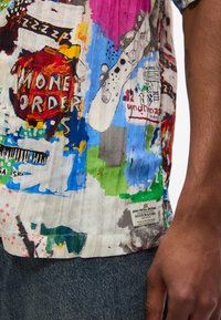 Ένα πολύχρωμο πουκάμισο με αφηρημένο σχέδιο, το οποίο φέρει το κείμενο «Money Order», πιτσιλιές από μπογιά και μια ετικέτα με την επιγραφή «Jean-Michel Basquiat Scotch & Soda» στο στήθος ενός ατόμου.