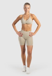 Soutien-gorge de sport beige avec des détails froncés et un short taille haute assorti, fabriqué en tissu texturé. Le mannequin se tient avec les mains sur les hanches.