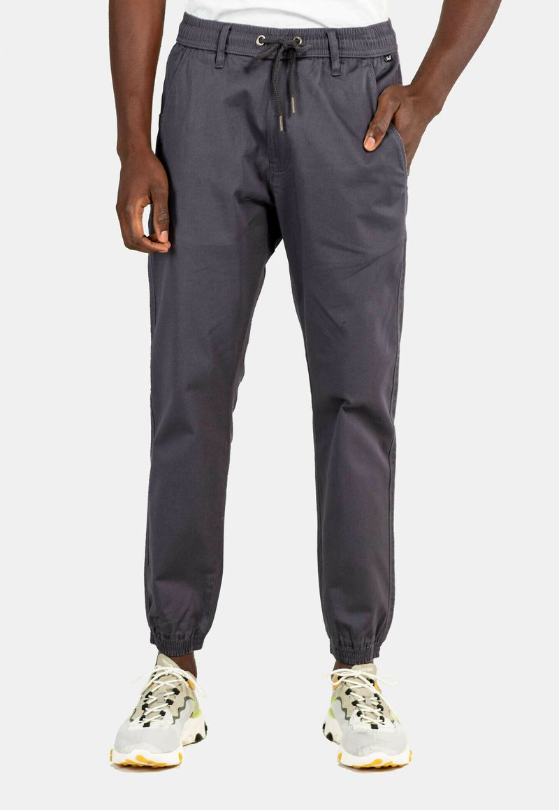 Pantalones jogger de color gris oscuro con cintura elástica, cordón, bolsillos laterales y puños ajustados. Llevados con zapatillas de color claro.