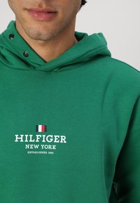 Zielona bluza z kapturem wykonana z miękkiego materiału, z kapturem na sznurki, bocznymi zatrzaskami oraz białym logo z małym detalem flagi na klatce piersiowej.