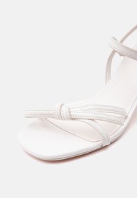 Λευκό σανδάλι ανοιχτού δαχτύλου με λεπτές ραμμένες λουρίδες και μια μικρή λεπτομέρεια κόμπου κοντά στο μπροστινό μέρος, σε απλό φωτεινό φόντο.