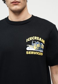 Άνδρας που φοράει μαύρο μπλουζάκι με κίτρινο και μπλε γραφικό εκχιονιστικού και το κείμενο "ICECREAM SERVICES" στο αριστερό στήθος.
