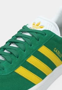 Πράσινο σουέντ αθλητικό παπούτσι με τρεις κίτρινες ρίγες, λευκή λαστιχένια σόλα και λευκή γλώσσα με κίτρινο λογότυπο. Εμφανίζεται το όνομα μοντέλου "GAZELLE".