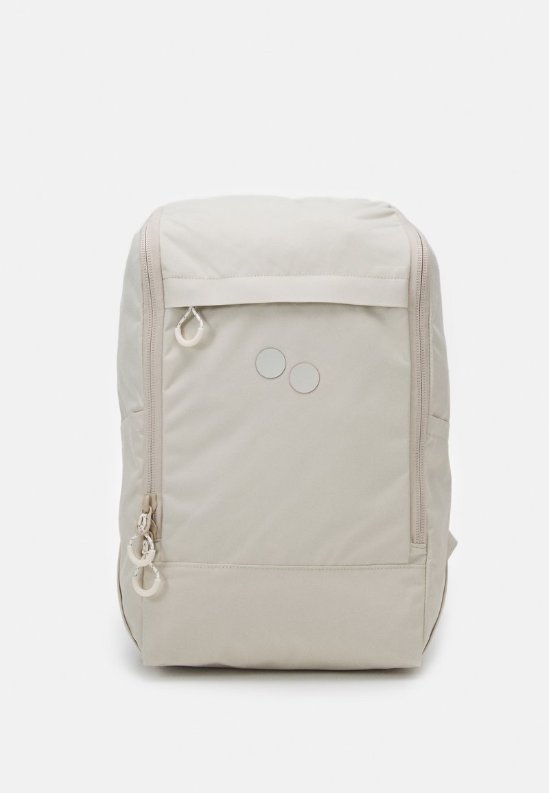 Sac à dos beige avec deux poches zippées à l'avant, des tirettes de fermeture éclair blanches et deux petits accents ronds près du centre supérieur avant.
