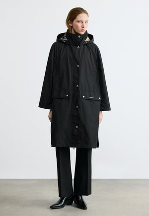 Naine, kes kannab pika musta kapuutsiga vihmamantlit, millel on klõpskangid, seisab hallil põrandal valge tausta ees.