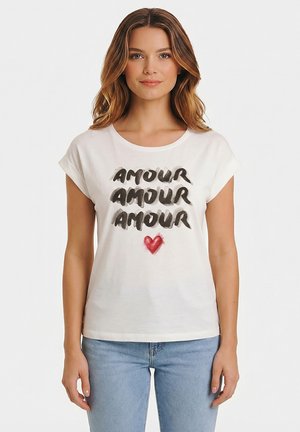 Donna che indossa una maglietta bianca con la scritta "AMOUR" ripetuta tre volte in corsivo nero e un cuore rosso sotto, abbinata a jeans azzurri.
