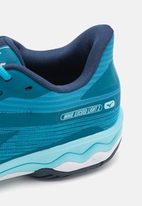 Scarpa sportiva in verde scuro e azzurro chiaro con tomaia in rete, colletto imbottito, design a onde sulla suola e logo del marchio visibile.