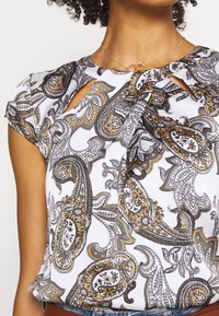 Blusa blanca con un patrón de paisley en dorado, negro y gris. Presenta un cuello con abertura y mangas cortas, fabricada en un material ligero.