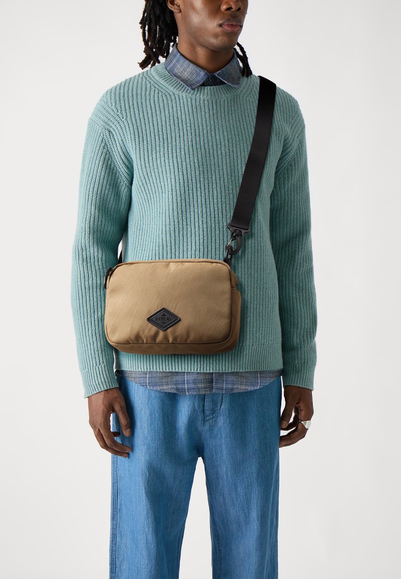Borsa a spalla in tela beige con logo nero a diamante, cinturino regolabile e superficie liscia, indossata sopra un maglione blu e una camicia a quadri.