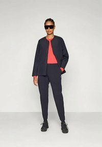 Giacca navy con collo rotondo sopra una maglia rossa brillante, abbinata a pantaloni navy e sneakers nere. Occhiali da sole eleganti completano il look.