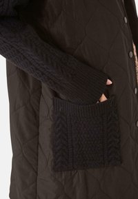 Tejido exterior negro acolchado con una manga de suéter acanalada y texturizada. Presenta un bolsillo de punto con patrón en la parte frontal, aumentando el interés táctil.