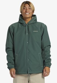 Veste à capuche verte avec fermeture éclair à l'avant, poignets élastiques et taille à cordon. Présente des coutures blanches et des poches latérales. Texture douce.