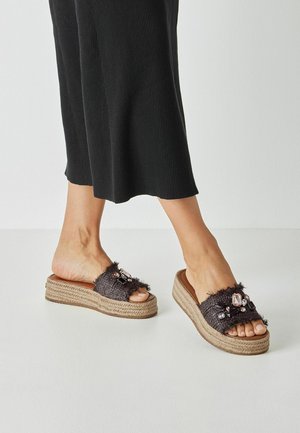 Fekete, texturált papucsok, puha pánttal, melyet kövek díszítenek, és fonott kötél talppal.