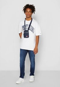 Maglietta bianca oversize con logo "TOMMY JEANS", jeans blu scuro e sneaker bianche. Piccola borsa a tracolla blu navy con cerniera e tracolla.