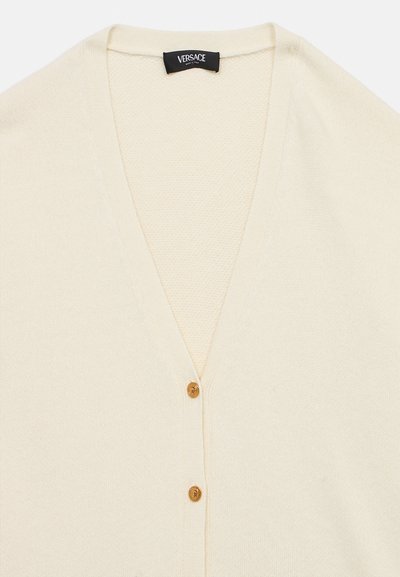 Κρεμ ζακέτα με σχέδιο V-λαιμό, διαθέτει υφή πλέξης και δύο χρυσά κουμπιά μπροστά. Η ετικέτα γράφει "Versace".