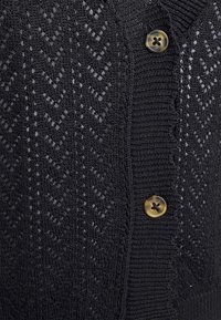 Svart cardigan med ett texturerat fiskbensmönster, med en slät yta och en rund, ljus sköldpaddsknapp.