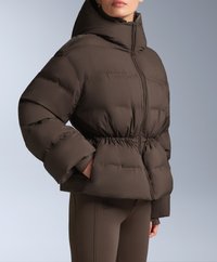 Hnedá puffermikina s vysokým golierom, stiahnutým pásom a nadýchanými rukávmi. Materiál sa zdá byť hladký s prešívanou textúrou. Zapínanie na zips.