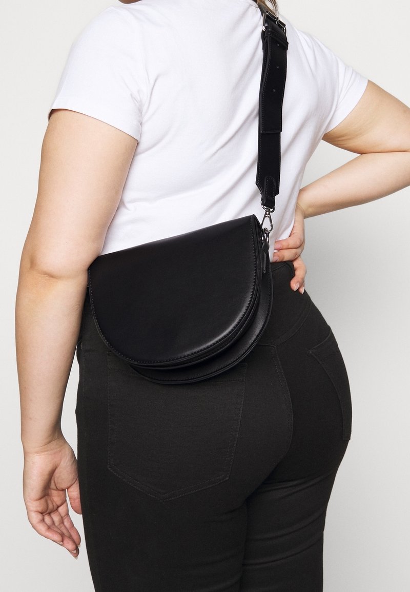 Černá kožená crossbody taška s designem zakřiveného klopa, nastavitelným popruhem a hladkým povrchem. Nositelná na těle přes bílou košili.