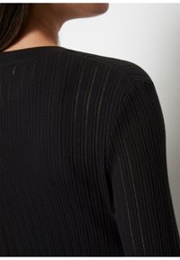 Czarny prążkowany dzianinowy element garderoby z pionowymi paskami i luźnym krojem. Tekstura wydaje się miękka, z lekką przejrzystością w niektórych miejscach.