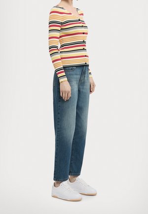 Personne portant un cardigan à rayures multicolores boutonné, un jean bleu et des baskets blanches, debout devant un fond uni.