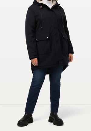Personne portant un parka noir à capuche avec grandes poches avant, jean bleu foncé et bottes noires épaisses, debout sur un fond blanc uni.