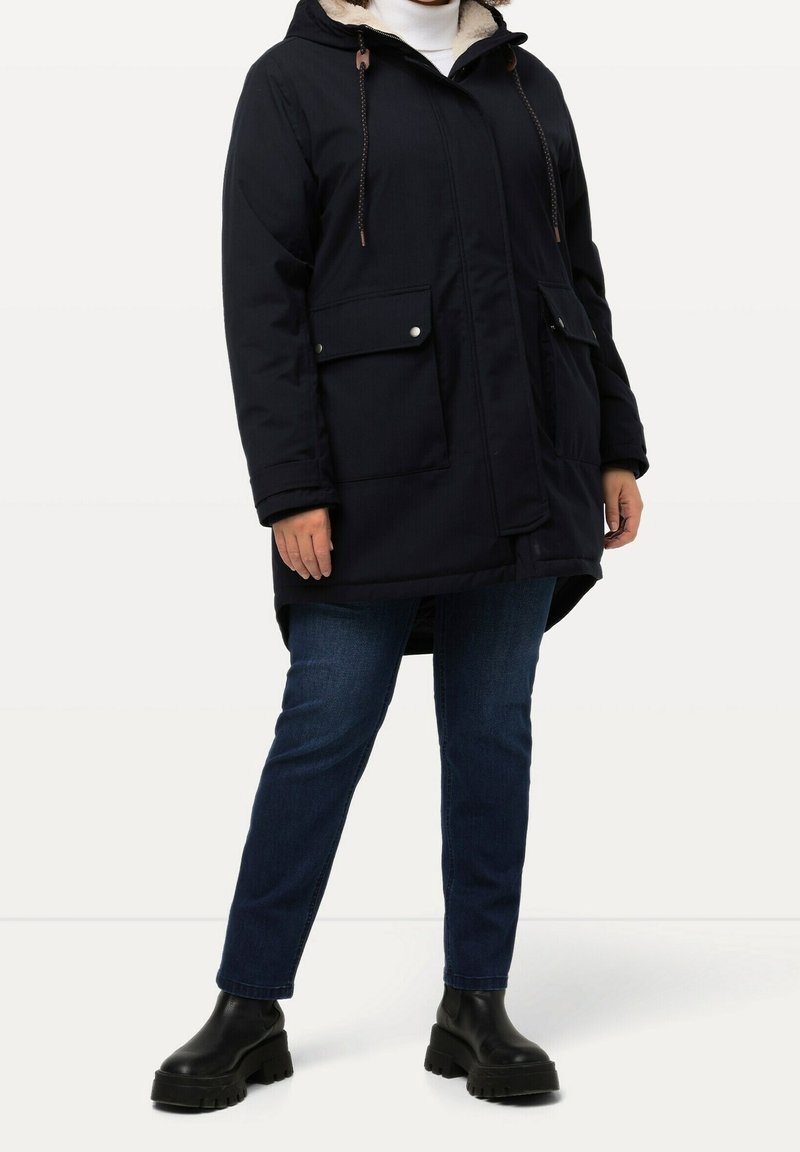 Personne portant un parka noir à capuche avec grandes poches avant, jean bleu foncé et bottes noires épaisses, debout sur un fond blanc uni.