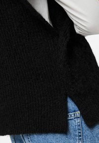 Svart stickad väst med en texturerad väv och en något oversized design, bärs över en ljusgrå långärmad skjorta, tillsammans med blå denim.