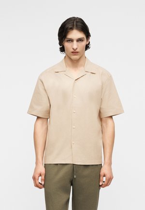 Νεαρός με κοντά, κυματιστά σκουρόχρωμα μαλλιά, που φοράει μπεζ κοντομάνικο πουκάμισο με κουμπιά και χακί παντελόνι, στέκεται μπροστά σε λευκό φόντο.