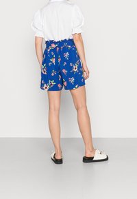 Modré květinové šortky s elastickým pasem a volánkovými detaily, zdobené živými růžovými a zelenými květinovými vzory. Nošeny s bílými sandály.