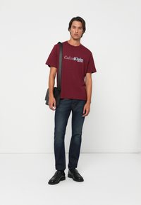 Man wearing a burgundy Calvin Klein t-shirt, dark jeans, black leather shoes, and carrying a black shoulder bag against a white background.