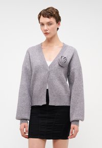 Harmaa neulottu neuletakki syvällä V-päällä, ribatulla hihalla ja ruusukoristeella vasemmalla puolella. Materiaali näyttää pehmeältä ja teksturoidulta.