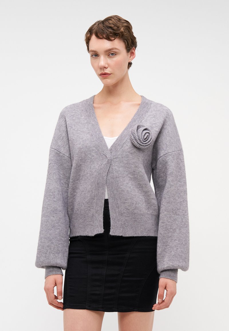 Harmaa neulottu neuletakki syvällä V-päällä, ribatulla hihalla ja ruusukoristeella vasemmalla puolella. Materiaali näyttää pehmeältä ja teksturoidulta.