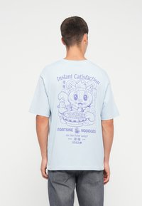 Man in een lichtblauw t-shirt met een cartoonkat die noedels eet en de tekst "Instant Catisfaction, Fortune Noodles, Are You Feline Lucky?" op de achterkant.