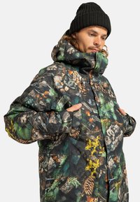 Mężczyzna w czarnej dzianinowej czapce noszący kurtkę w moro z nadrukiem szyszek, liści i gałęzi, z rękami w kieszeniach kurtki, patrzący w dół.