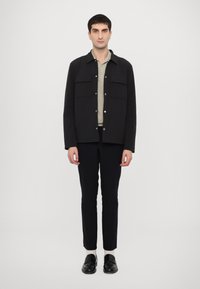 Jachetă neagră cu nasturi, cu două buzunare pe piept, purtată peste un top bej deschis, asociată cu pantaloni slim-fit negri și mocasini negri. Fundal simplu.