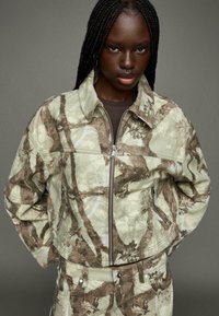 Camo-patroonjacket in lichtgroen en bruin, met een ritssluiting aan de voorkant, zijzakken en een cropped ontwerp. Gemaakt van textuur katoen.