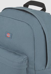 Mochila de tela azul con un acabado texturizado, un bolsillo frontal y un asa superior para llevar. Presenta un logo de marca multicolor.
