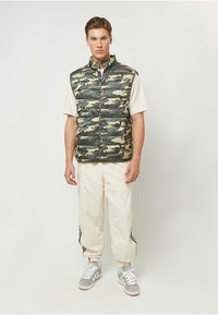 Camouflage Puffer-Weste mit hohem Kragen, sichtbarem Reißverschluss und gestepptem Muster. Über einem leichten Hemd getragen und mit cremefarbenen Jogginghosen und grauen Sneakers kombiniert.