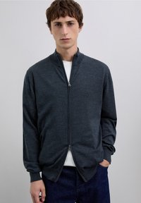 Dunkelgraue Strickjacke mit Reißverschluss aus weichem Material, mit Stehkragen und langen Ärmeln, getragen über einem weißen T-Shirt und blauen Jeans.