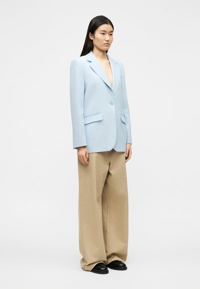 Γυναίκα που στέκεται φορώντας ένα γαλάζιο σακάκι με ένα κουμπί, μπεζ παντελόνι φαρδιάς γραμμής με μικρές τρούκ, και μαύρα παπούτσια σε ένα απλό λευκό φόντο.