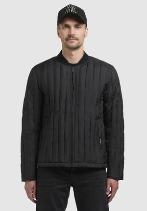 Schwarze gesteppte Jacke mit vertikaler Naht und einem Frontreißverschluss. Gerippter Kragen und seitliche Taschen. Das Model trägt eine schwarze Mütze dazu.